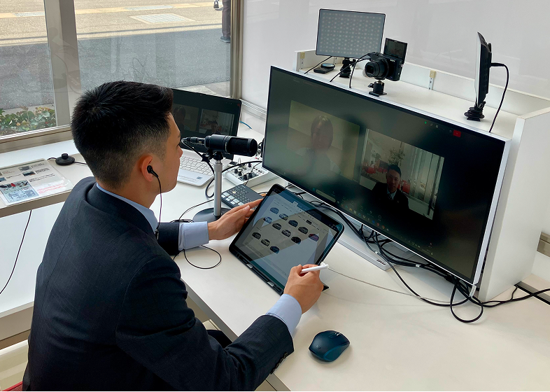 スーツ姿の男性がデスクでタブレットを操作しながら、モニターとカメラを使ってオンライン対応しているイメージ写真